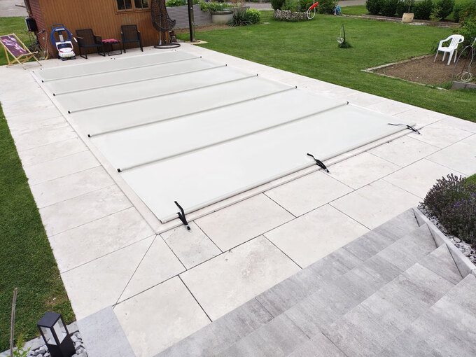 Lichtgraue Poolabdeckung auf einer modernen Stein-Terrasse mit Garten und Holzhaus im Hintergrund.