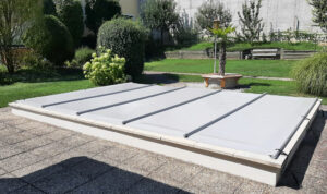 Lichtgraue Poolabdeckung auf erhöhtem Becken in einem gepflegten Garten.