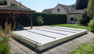 Lichtgraue Poolabdeckung auf einem erhöhten Becken in einem gepflegten Garten mit Holzpavillon und Grillplatz.