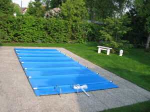 Hellblaue Poolabdeckung auf einem eingelassenen Pool in einem gepflegten Garten mit grüner Rasenfläche und Sitzbank.