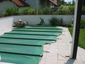 Dunkelgrüne Poolabdeckung auf einem eingelassenen Pool mit geschwungener Form, befestigt mit Spannriemen in einem gepflegten Garten mit Sichtschutzwand.